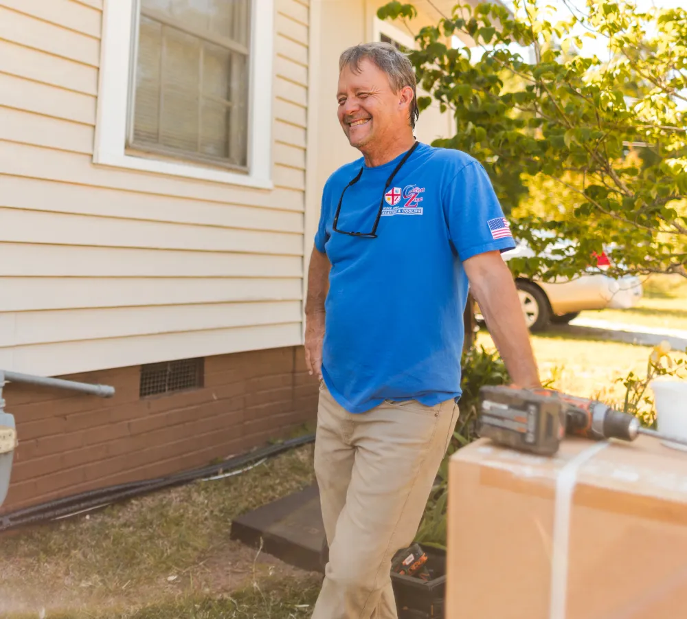 Smiling man in blue T-shirt stands outdoors by a house, near a box and power tool, on a sunny day.