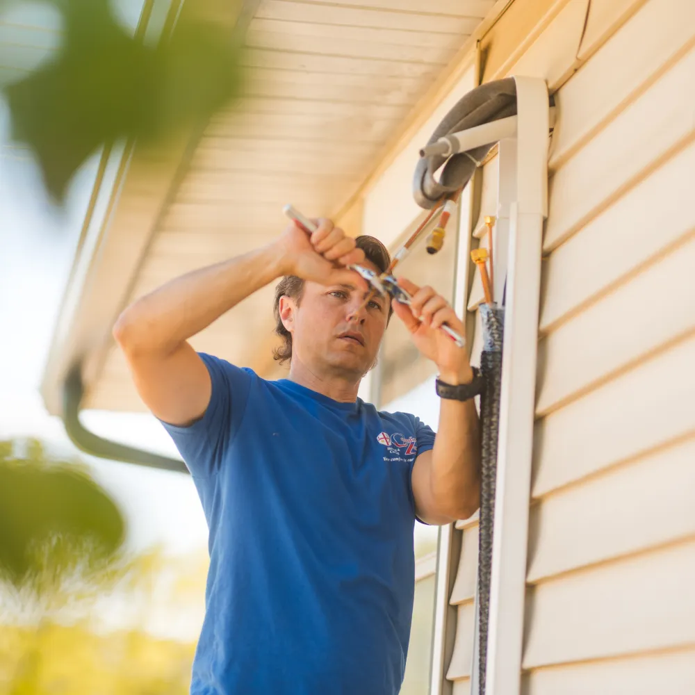 Man in blue shirt using tools to work on pipes outside a house with beige siding.