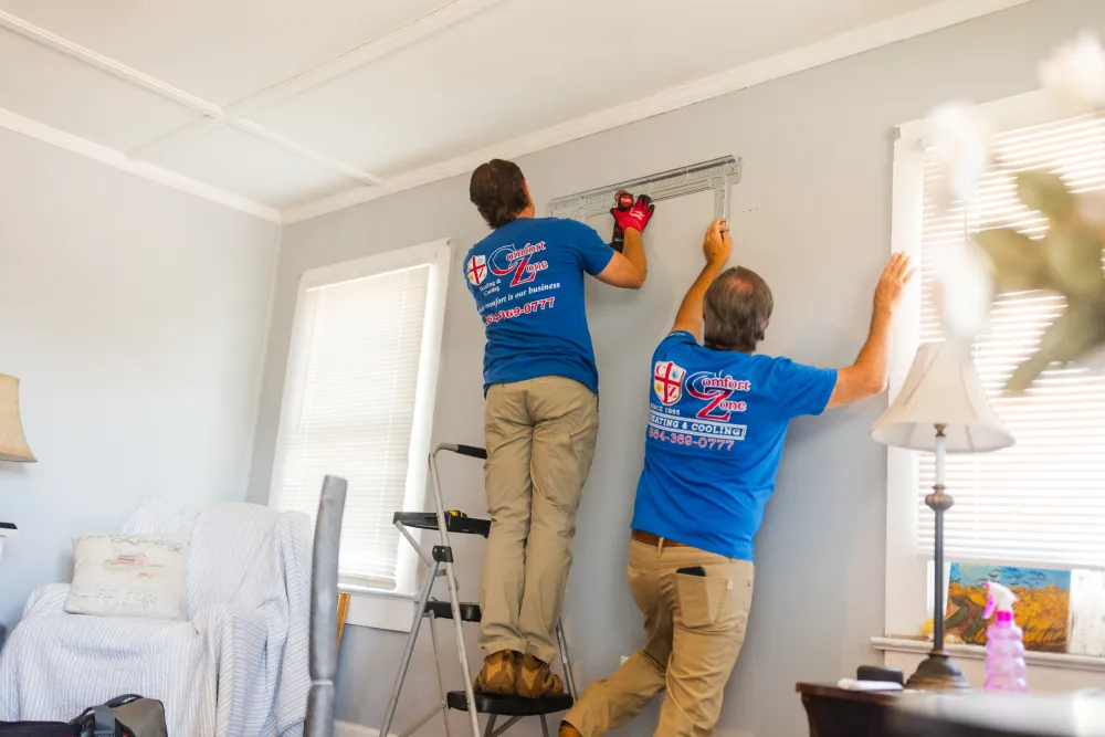Two people in blue shirts install window blinds in a living room. One stands on a step stool.