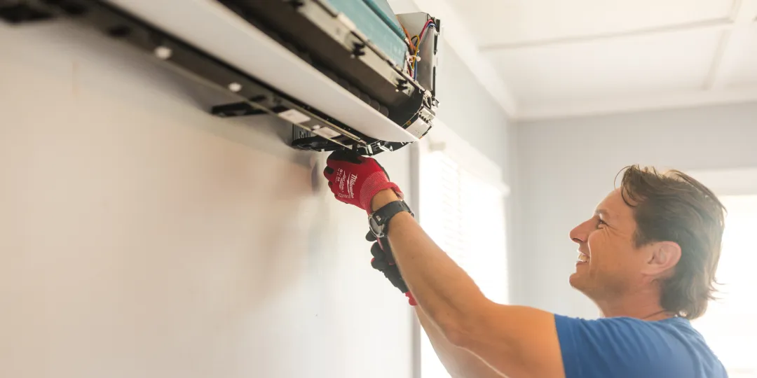 Smiling man in gloves repairs a wall-mounted air conditioner unit with a screwdriver.