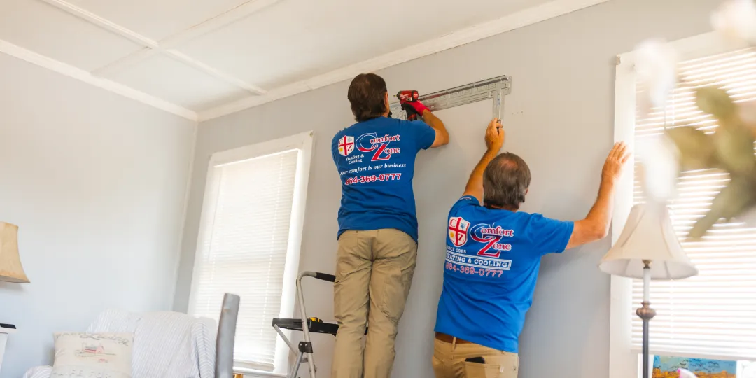 Two workers in blue shirts install a large window blind on a living room wall using tools and a ladder.