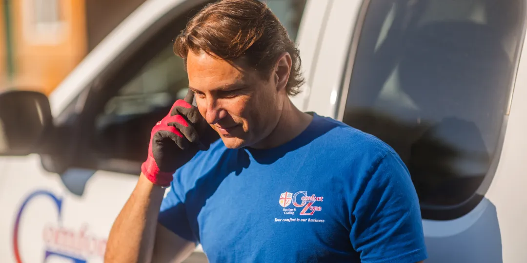 Man in blue shirt and work gloves talks on a cell phone beside a white service van.