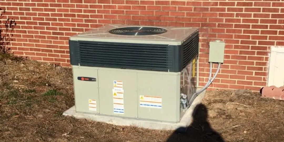Outdoor air conditioning unit next to a brick wall on a concrete pad, with electrical box attached.