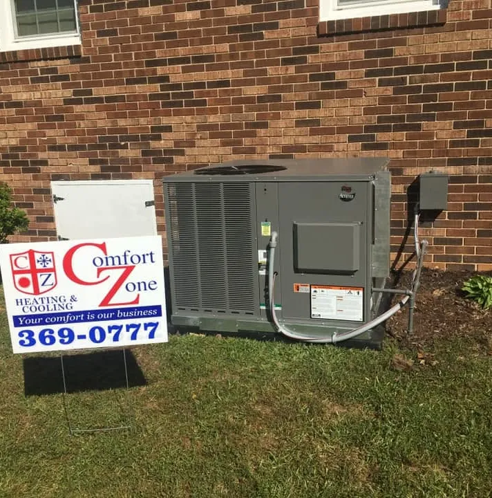 Outdoor HVAC unit installed next to a brick building, with a Comfort Zone service sign on the lawn.