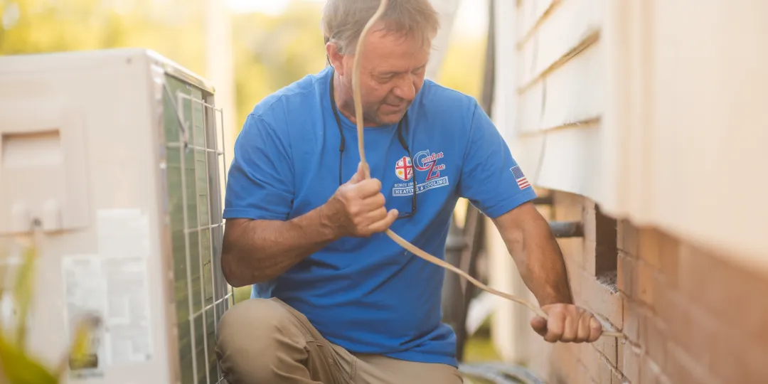 Man in blue shirt repairing outdoor air conditioning unit next to a house on a sunny day.