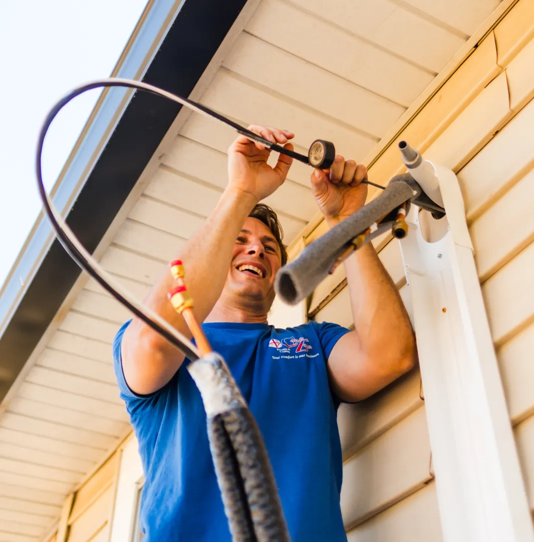 Man in blue shirt installing or repairing an air conditioning unit on the exterior wall of a house.