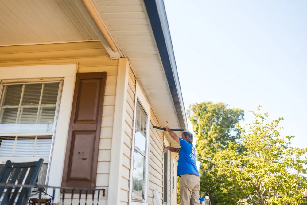 A person in a blue shirt paints trim on the side of a tan house under a clear sky.